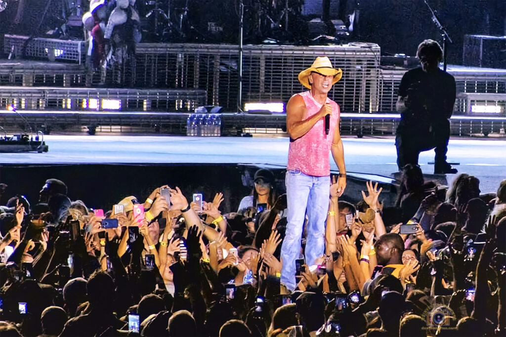 A close-up shot of country star Kenny Chesney standing at the edge of the stage at the Rose Bowl, wearing his signature straw cowboy hat and a pink sleeveless shirt. He is holding a microphone and smiling while hundreds of fans in the front row reach their hands up and hold up cell phones to capture the moment.