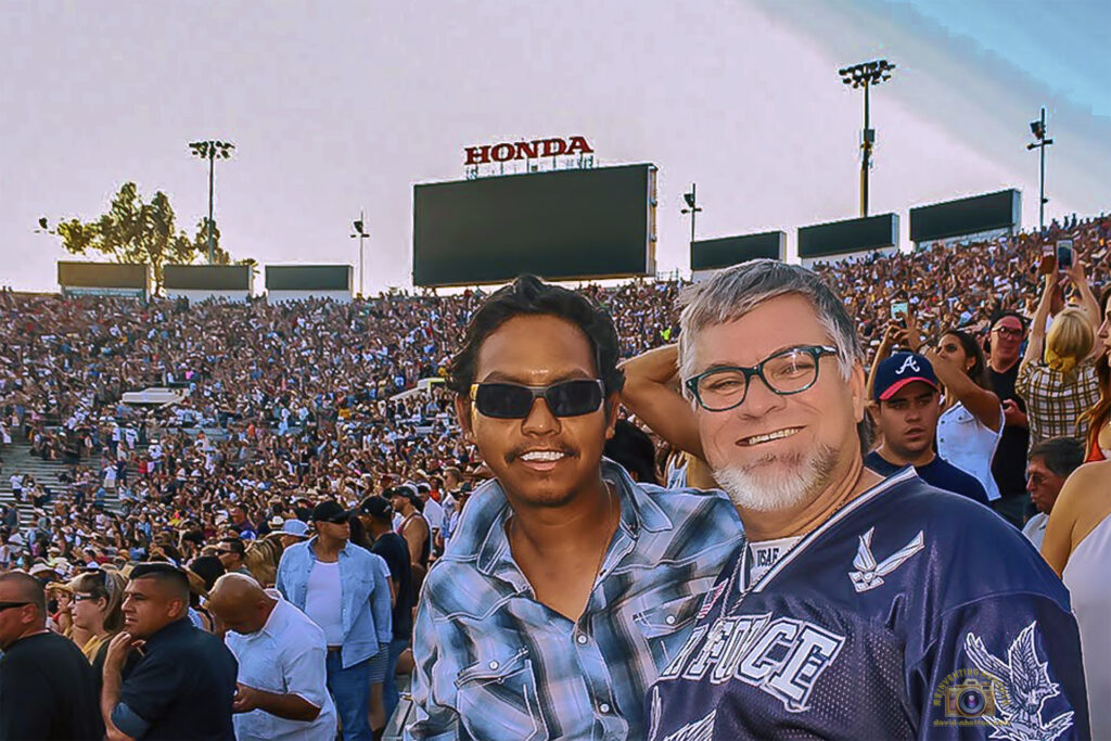A selfie of me and my husband, Gio, smiling in the sunny stands at the Rose Bowl Stadium. Gio is wearing a US Air Force-style jersey. The background is filled with a massive crowd and the stage setup for the Kenny Chesney Big Revival Tour.