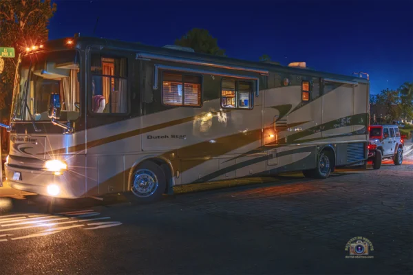 A tan and green Newmar Dutch Star Class A motorhome parked at night with headlights on, flat-towing a silver Jeep Wrangler "toad" behind it.