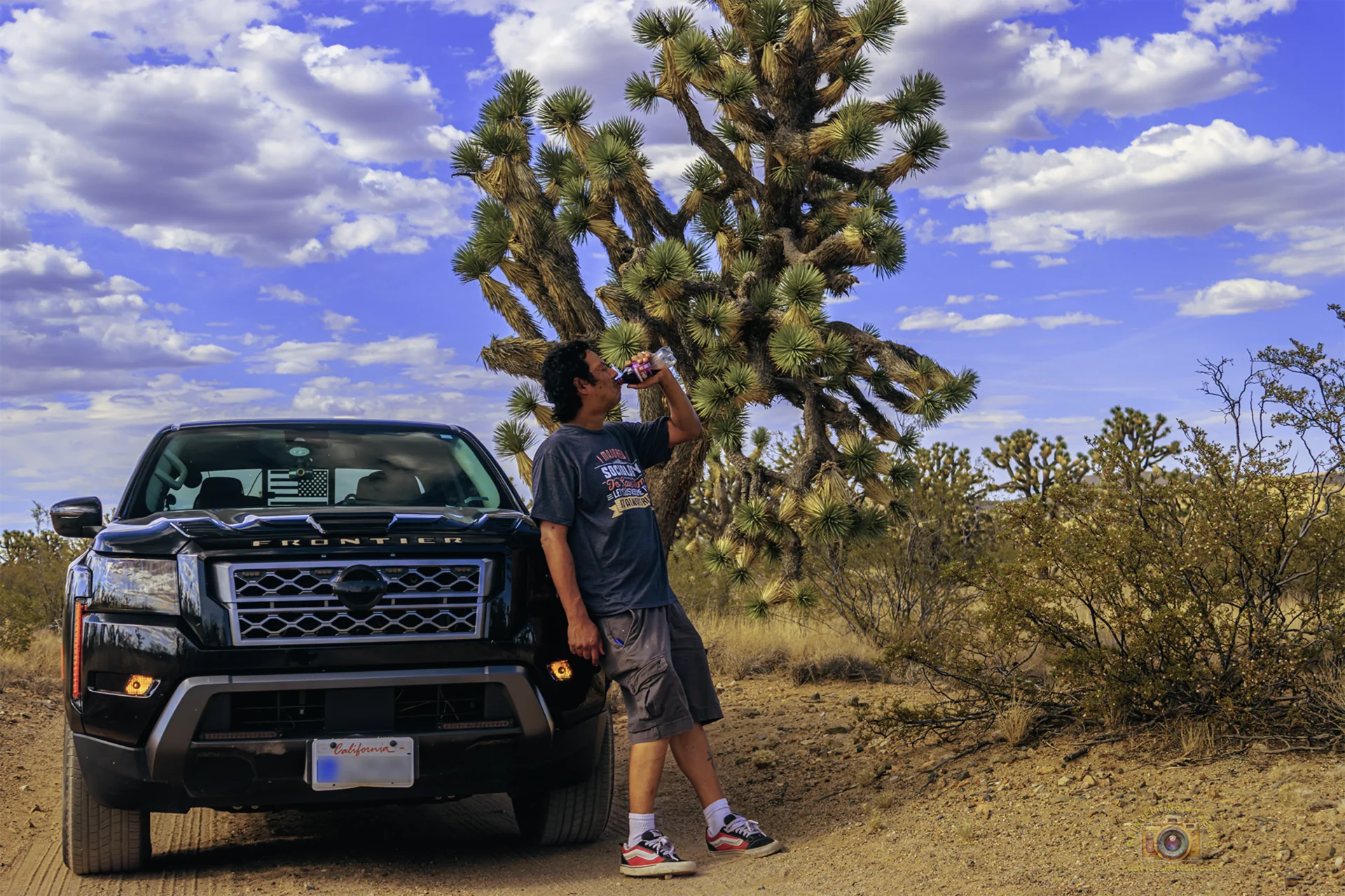 Gio drinking from a can while leaning against a black Nissan Frontier truck on a desert dirt road in the Ancient Ones Joshua Tree Forest of Nevada, beneath a large Joshua tree and a blue sky with white clouds.