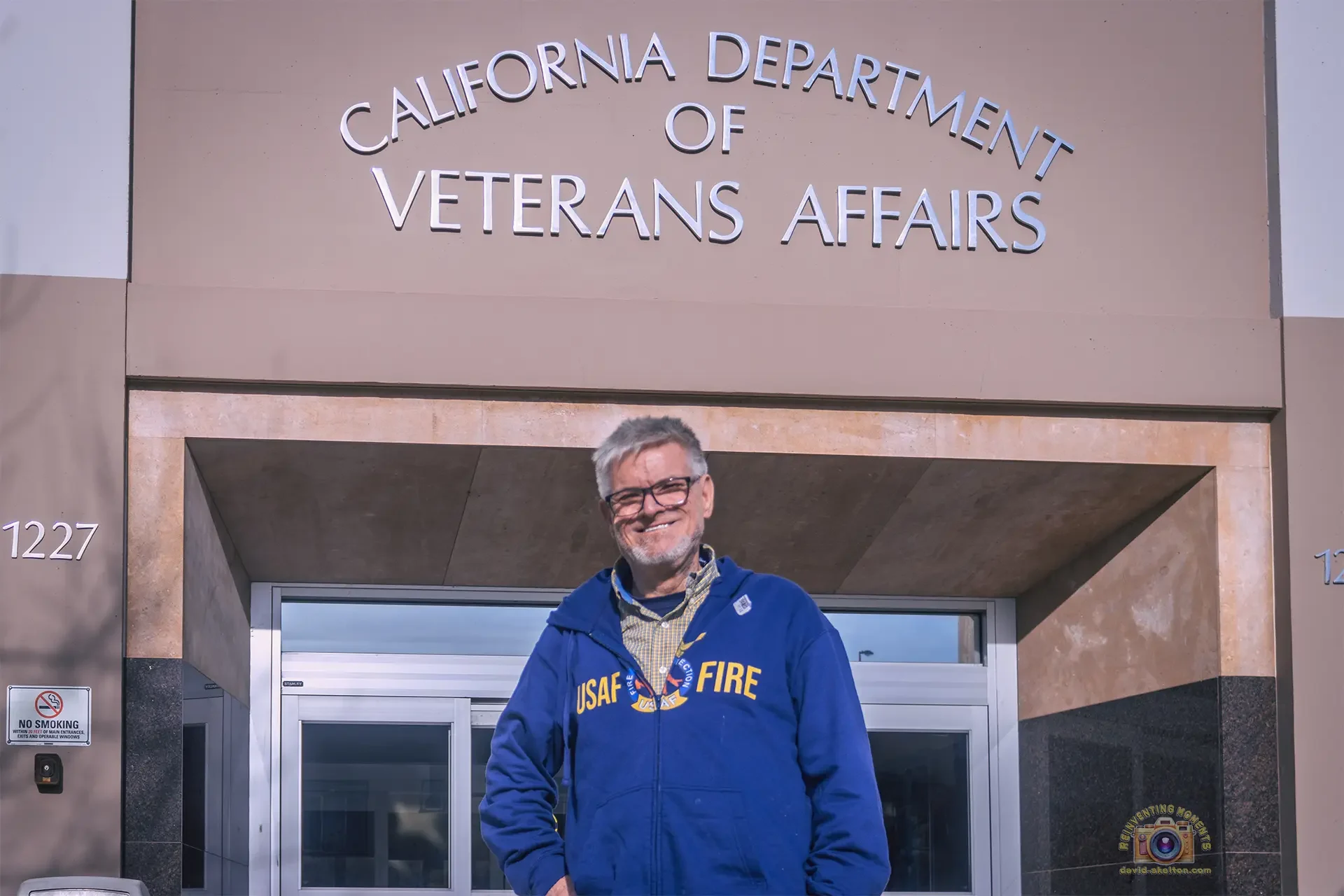 A smiling man wearing a blue USAF Fire Protection hoodie standing in front of the main entrance to the California Department of Veterans Affairs Headquarters building in Sacramento.