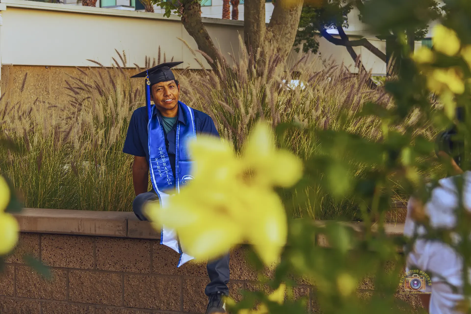 A shallow depth of field portrait of a male graduate named Gio wearing a blue CSUSB sash and graduation cap, framed by a blurred yellow rose in the foreground.