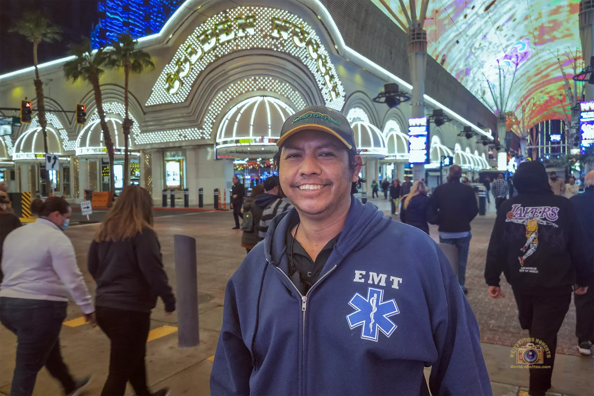 Gio smiling on Fremont Street in Downtown Las Vegas at night, wearing a Crafton Hills College hat and a blue EMT hoodie, with the brightly lit Golden Nugget casino facade and neon canopy overhead.