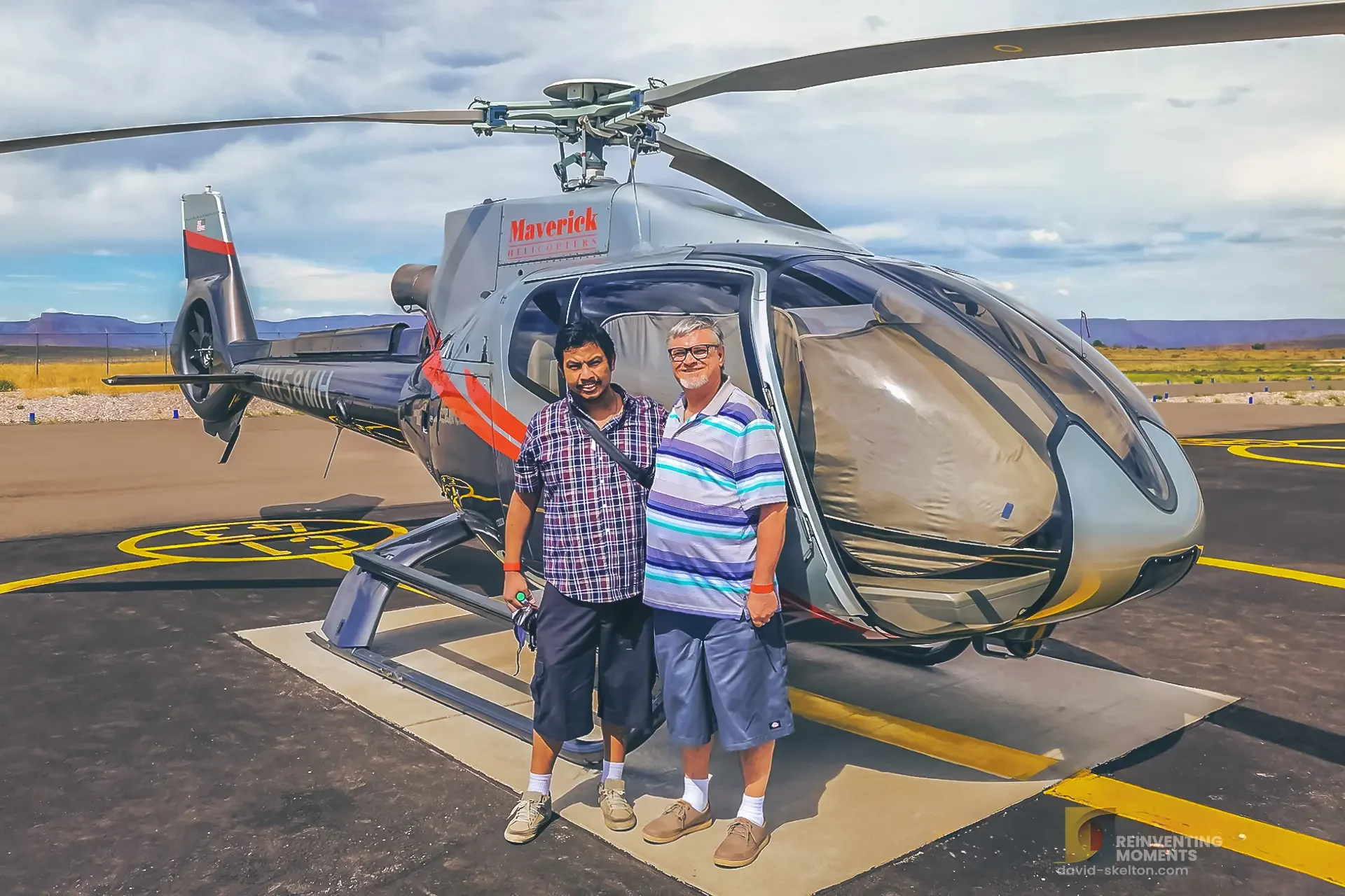 David and Gio standing proudly together on the tarmac in front of a grey and orange Maverick Helicopters Airbus EC130 at the Grand Canyon West heliport.