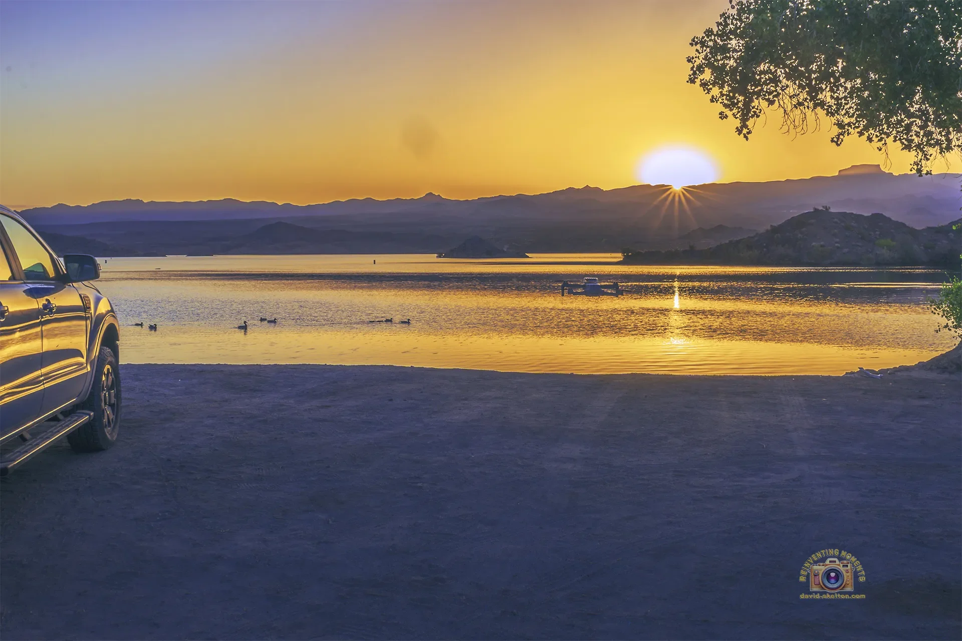 A blue Ford Ranger parked on the dirt shore of Nevada Telephone Cove at sunrise, with a DJI Mini 4K drone hovering over the golden water of Lake Mohave as the sun peeks over the Black Mountains of Arizona