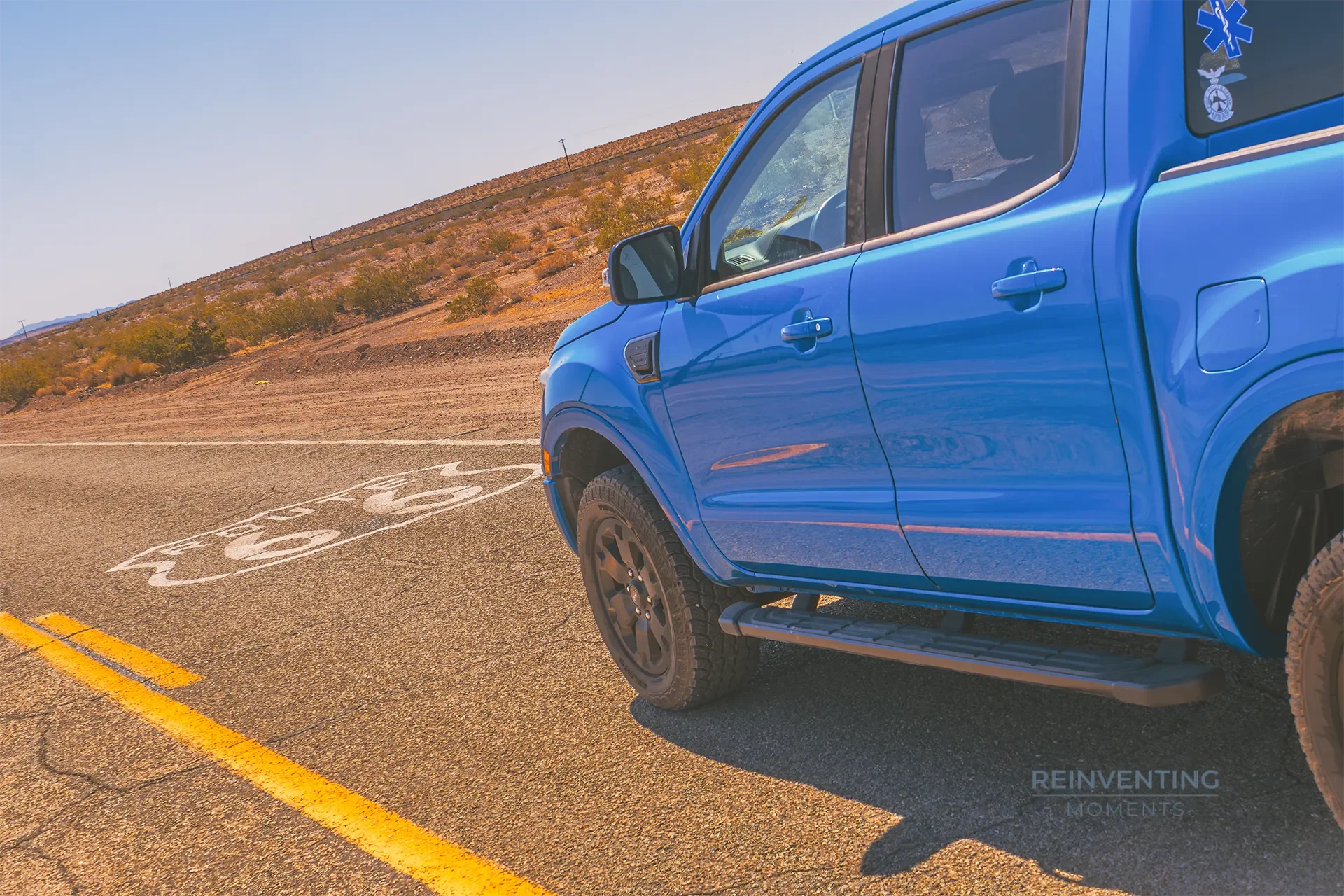 Side profile view of a blue Ford Ranger pickup truck parked over yellow lines on a historic desert highway under a clear blue sky.