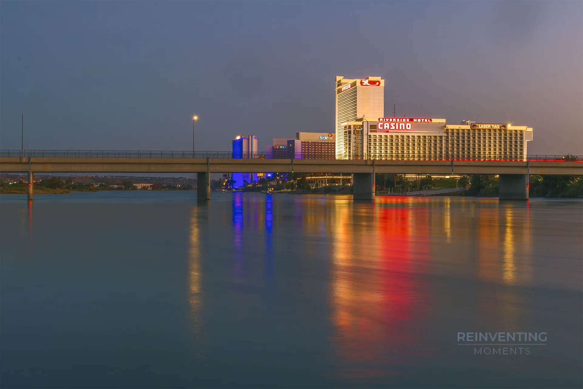 A long exposure night photograph of illuminated Laughlin casino towers, including the Riverside Resort, reflecting bright red and blue neon lights onto the calm Colorado River beneath a concrete bridge.