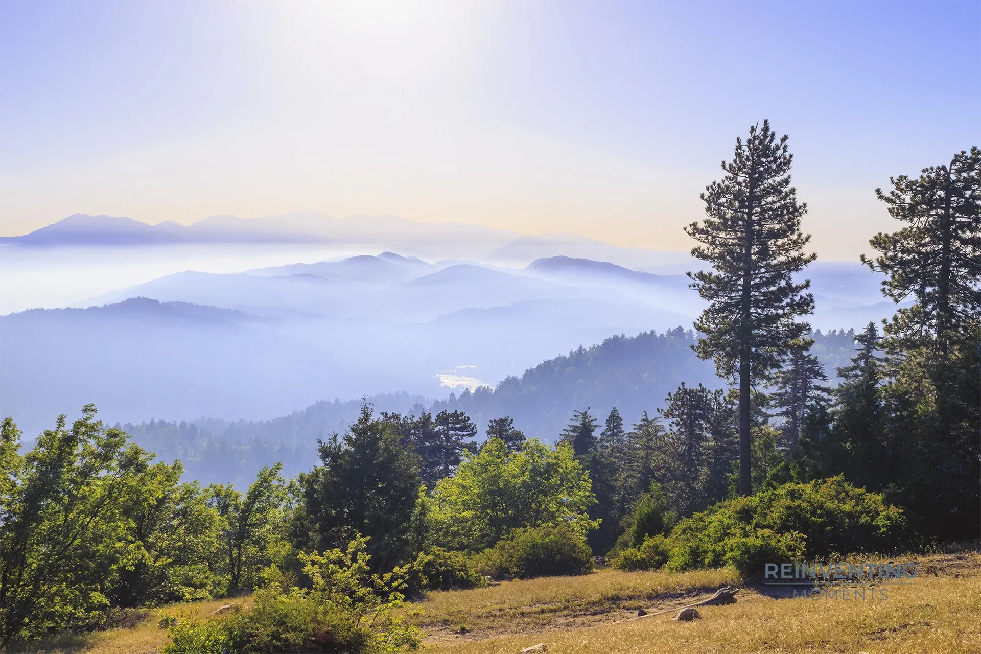 Mist-covered San Gabriel Mountains view from Strawberry Peak in Crestline, California, featuring Mt. Baldy and Lake Gregory under a hazy morning sky.