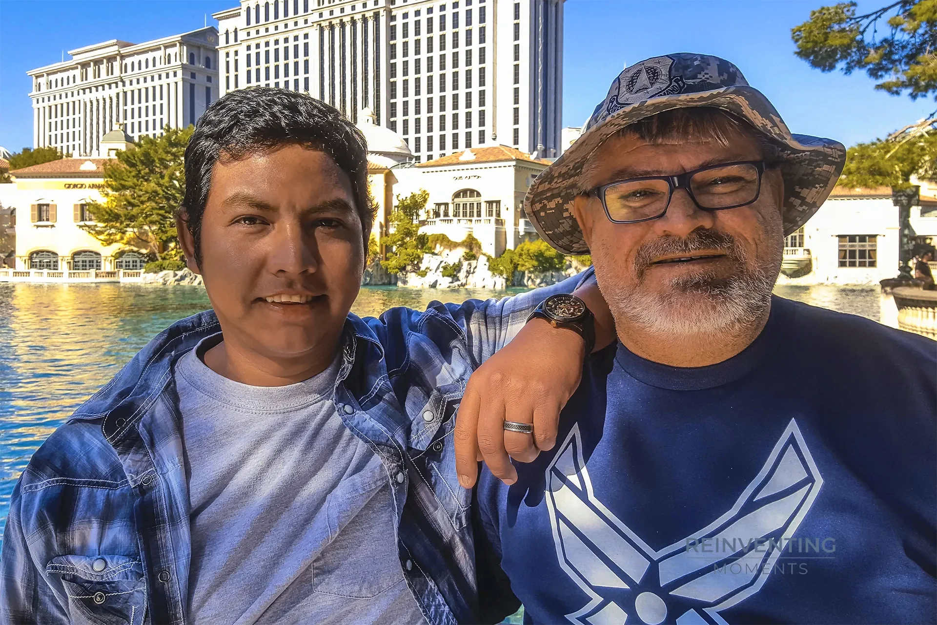 David and Gio smiling together in front of the blue waters of the Bellagio Fountains and Caesars Palace along the Las Vegas Strip.