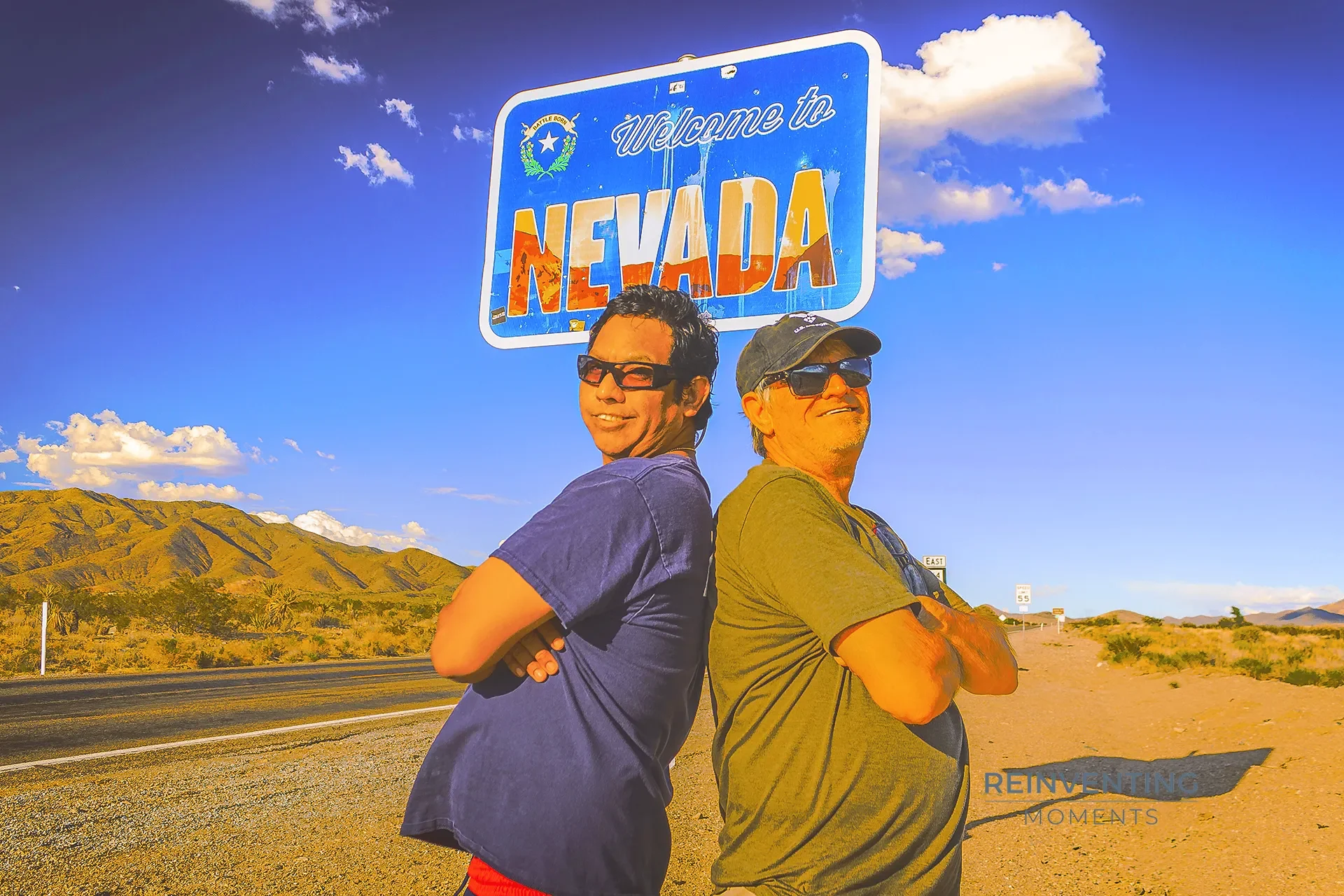 David and Gio Skelton smiling back-to-back under the Welcome to Nevada sign along a desert highway near the California border.