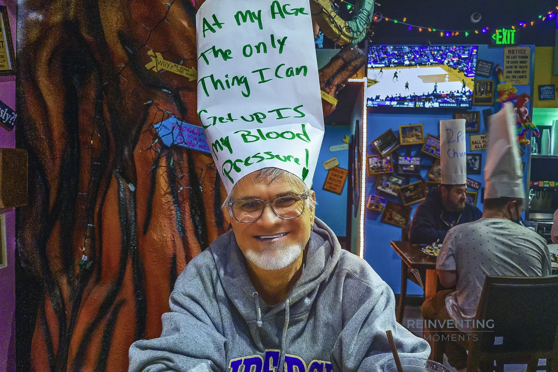 A man wearing a tall white paper hat with handwritten sarcastic text reading At my age the only thing I can get up is my blood pressure at Dick's Last Resort in Las Vegas.