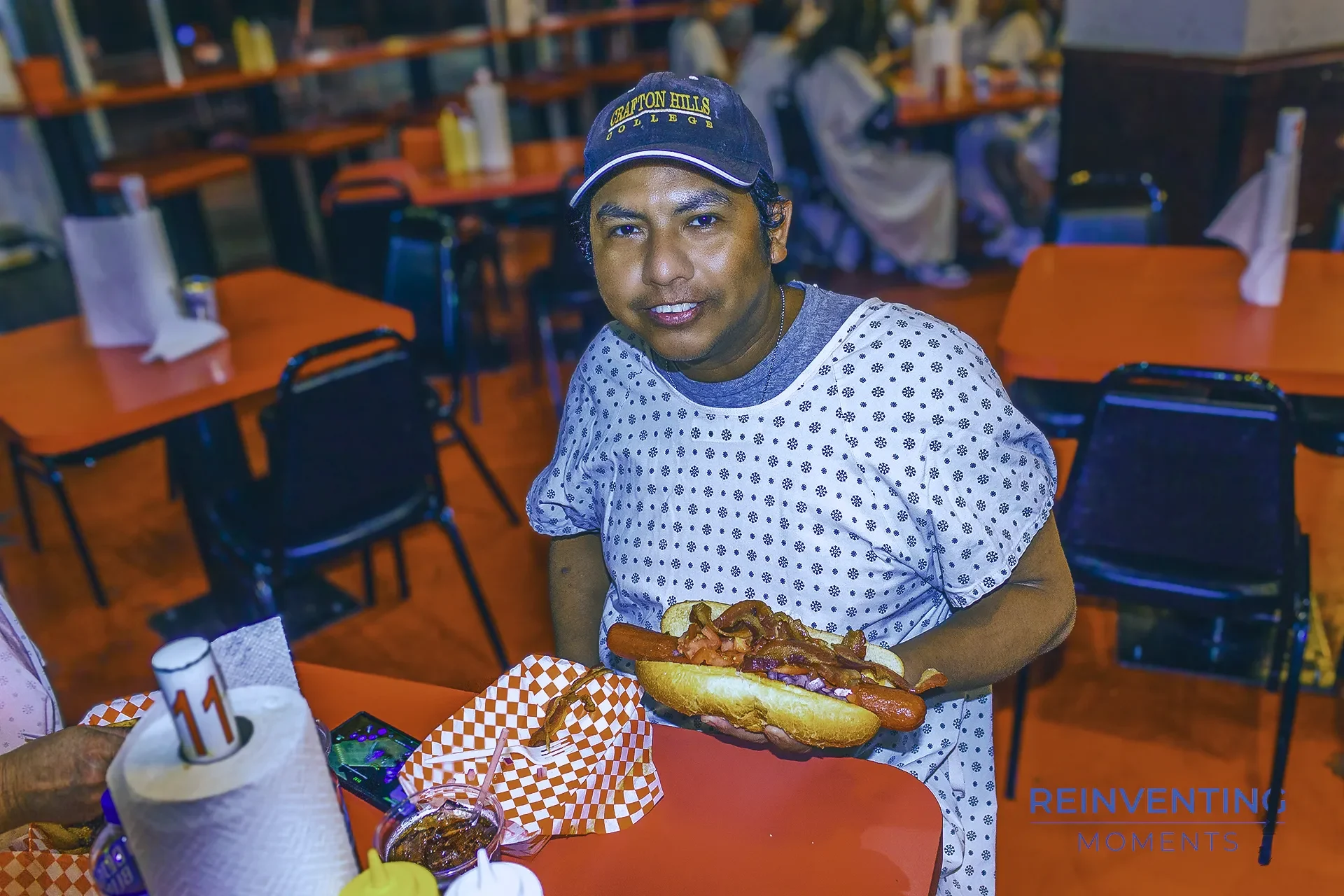 Gio sitting at a diner booth wearing a hospital gown holding a giant loaded hot dog at Heart Attack Grill in Las Vegas