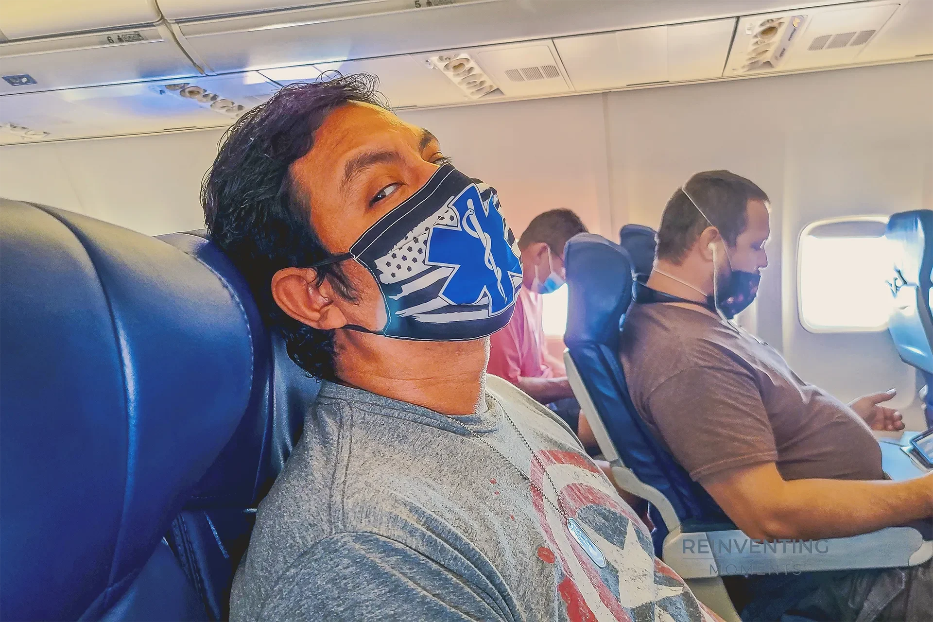 Passenger Gio wearing an EMS graphic face mask seated in an airplane cabin on a flight from Ontario to Las Vegas