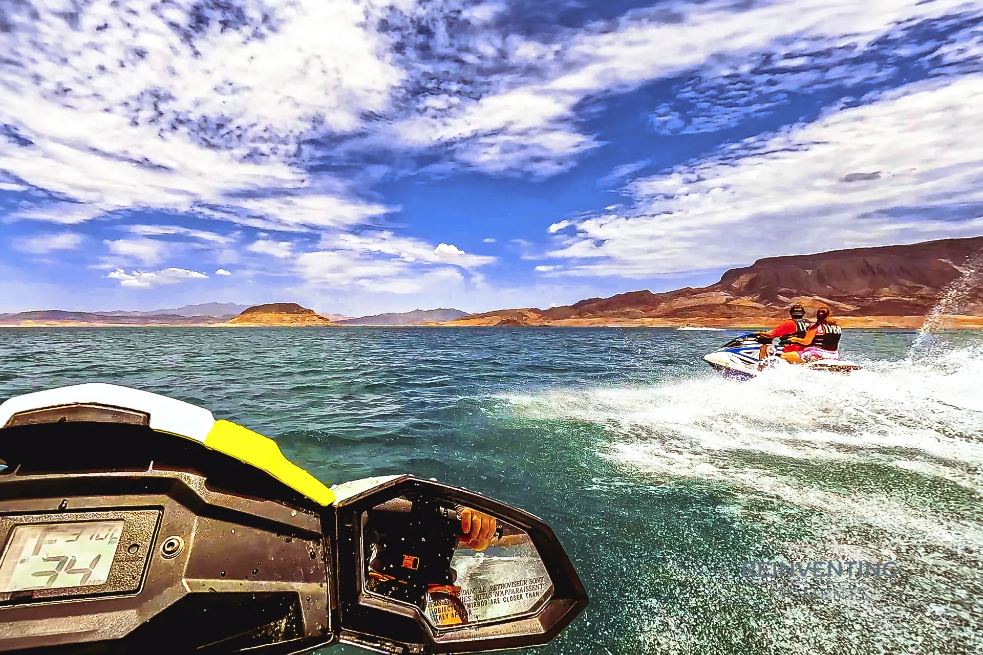 POV view from a jet ski handlebar showing another jet ski speeding across blue water on Lake Mead toward distant desert mountains with white bathtub rings.